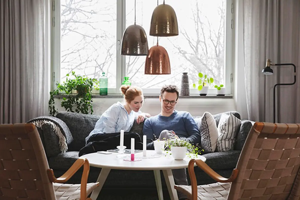 Een stel zit lachend op de bank en bekijken iets op hun mobiel.
